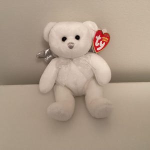 May include: White plush teddy bear with a silver glitter halo and wings. The bear has a red Ty Beanie Babies tag attached to its ear.