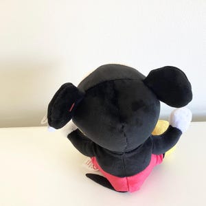 May include: A plush Mickey Mouse toy, viewed from the back. The toy features a black body, large black ears, white gloves, and red shorts. A yellow object is held in one hand. The toy is on a white surface.