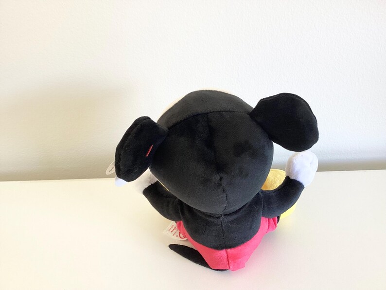 May include: A plush Mickey Mouse toy, viewed from the back. The toy features a black body, large black ears, white gloves, and red shorts. A yellow object is held in one hand. The toy is on a white surface.