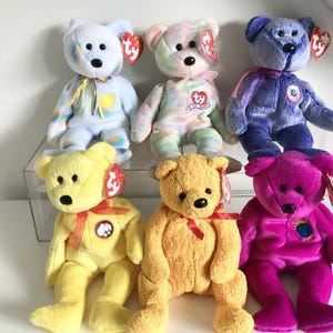 May include: Six colourful plush teddy bears with black button eyes and attached tags. The bears are in shades of blue, pink, yellow, and purple. Each bear has a ribbon around its neck. The bears are sitting on a white surface.