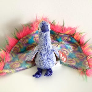May include: A purple and blue peacock plush toy with a colorful, iridescent tail. The tail is made of fluffy, multi-colored fur. The toy has a red and white tag attached to it.