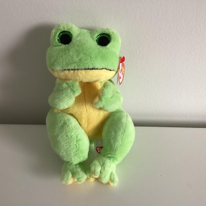 May include: A plush frog toy with a light green body and yellow belly. The frog has large, glittery green eyes and a red tag attached to its ear. The toy is sitting upright on a white surface.