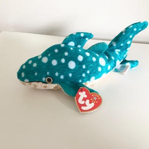 May include: A plush toy whale shark in teal with white spots. The stuffed animal has a white belly and a red heart-shaped tag attached. The toy is lying on a white surface.