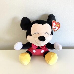 May include: A plush Mickey Mouse toy with black ears, a cream-colored face, and a red bow tie. The toy has a red body, yellow feet, and white gloves. A red heart-shaped tag is attached to the ear.