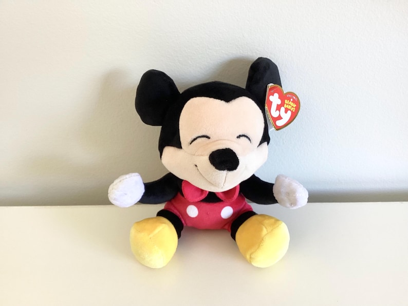 May include: A plush Mickey Mouse toy with black ears, a cream-colored face, and a red bow tie. The toy has a red body, yellow feet, and white gloves. A red heart-shaped tag is attached to the ear.