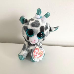 May include: A plush giraffe toy with a white and gray spotted pattern. It has large, sparkly blue eyes and glittery teal accents on its horns, ears, and feet. A heart-shaped tag is attached.