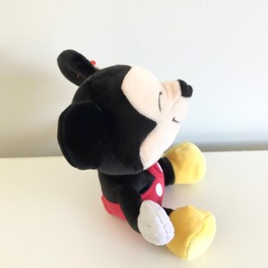 May include: A plush Mickey Mouse toy, featuring a black body, white face, and yellow gloves and shoes. The toy wears a red shorts with white buttons and a red bow. The toy is sitting on a white surface.