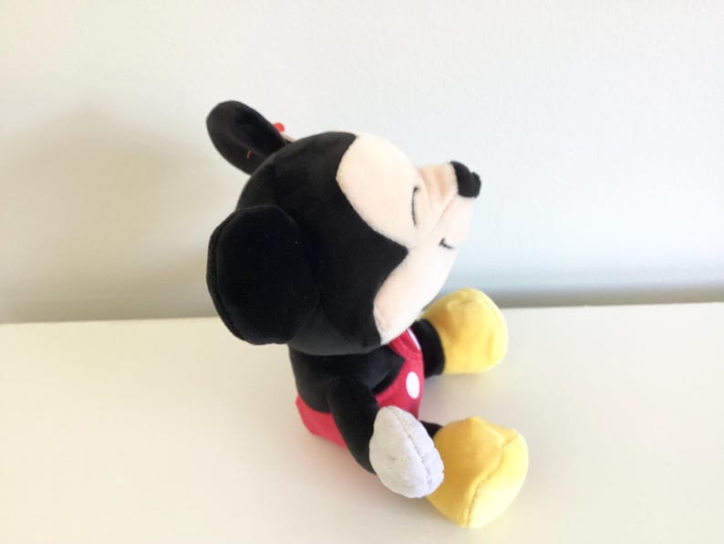 May include: A plush Mickey Mouse toy, featuring a black body, white face, and yellow gloves and shoes. The toy wears a red shorts with white buttons and a red bow. The toy is sitting on a white surface.