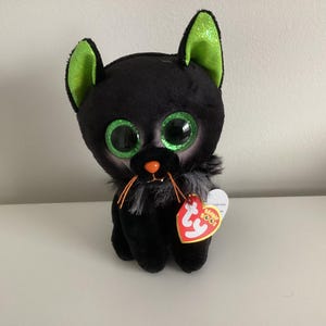 Peut inclure: Un jouet en peluche noir en forme de chat avec de grands yeux verts scintillants et des oreilles bordées de vert. Il a un nez et des moustaches oranges, avec un col en fourrure grise. Une étiquette rouge en forme de cœur est attachée.