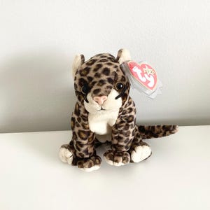 May include: A plush leopard toy with a spotted pattern in brown and tan. The stuffed animal has a white belly, paws, and ears, with a pink nose and large, dark eyes. A red tag is attached to the ear.