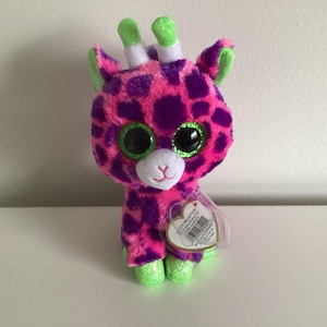 May include: Pink and purple giraffe plush toy with green horns and large, glittery eyes. The toy has a heart-shaped tag attached to it.