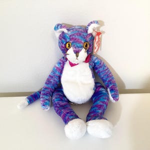 May include: A plush cat toy with a white chest and paws, and a pink bow. The cat's body is a mix of blue and purple colors. It has yellow eyes and a red tag.