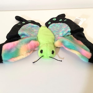 May include: A plush butterfly toy with black wings, rainbow-coloured accents, and a lime green body. The wings are spread out, and the toy has black eyes and antennae. A tag is attached.