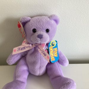 May include: Purple plush teddy bear with a pink ribbon that says "Happy Mother's Day". The bear has black eyes and a small pink nose. The bear is wearing a flower on its chest. The bear is holding a tag that says "Play Online!"