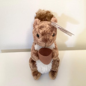 May include: A plush squirrel toy with brown fur, a white chest, and a fluffy tail. The squirrel is holding a brown acorn. A clear plastic tag is attached to the squirrel's head. The toy is on a white surface.