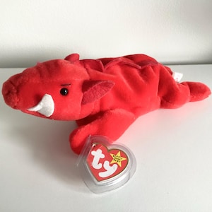 May include: A red plush dragon toy with white tusks and a black eye. The dragon has a tag attached to it. The toy is made of a soft material and is lying down.