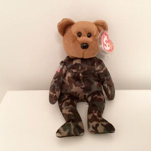 Könnte beinhalten: Ein brauner Plüschbär mit einem Tarnmuster-Uniform und einem kleinen amerikanischen Flaggenaufnäher. Der Bär hat eine braune Schnauze, schwarze Augen und ein rotes Etikett. Ein Sammlerspielzeug.