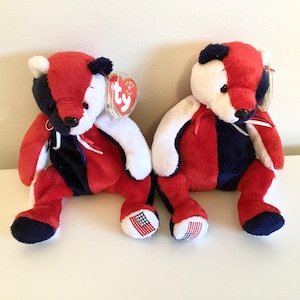 Op de afbeelding: Twee pluche teddyberen in rood, wit en blauw, met een Amerikaanse vlag op de voet. Elke beer heeft een rode kop, een wit gezicht en armen, en een blauw en rood lichaam. Ze hebben een TY-label.