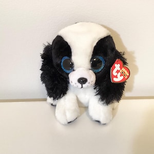 May include: A black and white plush dog toy with large blue glittery eyes and a red heart-shaped tag. The soft toy is positioned on a white surface.