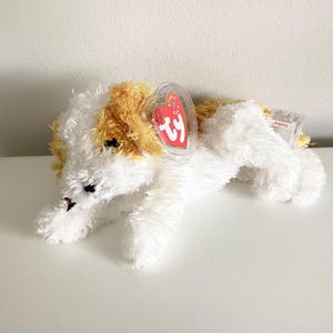 May include: A soft toy dog with white and tan fur. The toy has black eyes and a red heart-shaped tag attached to its ear. The dog is lying down on a white surface.