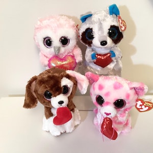 May include: Four plush animal toys: an owl, a raccoon, a dog, and a cat. The owl is pink with a heart, the raccoon is white and gray with a heart, the dog is brown and white with a heart, and the cat is pink with spots and a heart.