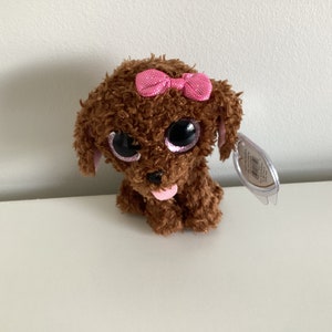 May include: A brown plush puppy with large, glittery eyes and a pink bow. The toy dog has a pink tongue sticking out and a tag attached. The plush toy is sitting on a white surface.