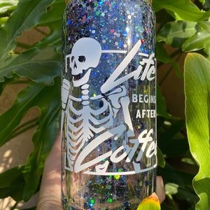 May include: A clear glass tumbler with a bamboo lid and a straw. The tumbler is filled with glitter and features a white skeleton graphic holding a coffee cup. Text on the tumbler reads "Life Begins After Coffee."