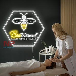 Könnte beinhalten: Ein hexagonales Neonschild mit einer weißen Bienengrafik mit gelbem Hinterleib, das den Text BeeWaxed WAX & SKINCARE STUDIO in weißen und gelben Neonröhren zeigt. Das Schild ist an einer dunklen Wand über einer Behandlungsliege montiert.