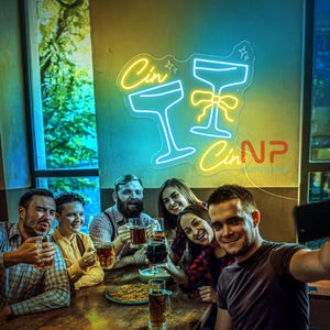 Pu&ograve; includere: Un'insegna al neon gialla e blu con la scritta "Cin" e due calici da cocktail. Un gruppo di persone &egrave; riunito attorno a un tavolo, tiene in mano delle bevande e si fa un selfie.