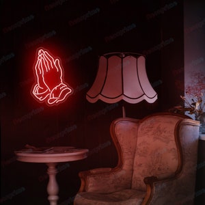 May include: A red neon sign depicting a pair of hands in a praying position. The sign is mounted on a wall in a room with a lamp and a chair.