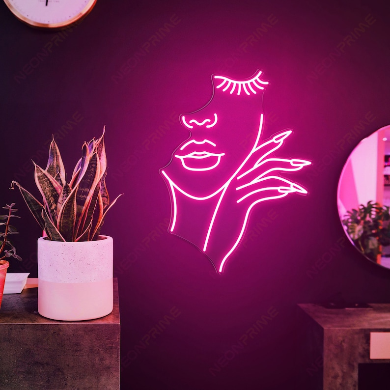 May include: Pink neon sign depicting a stylised outline of a woman's face with eyelashes, lips, and a hand with manicured nails.