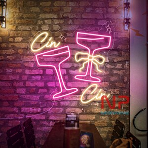 Pu&ograve; includere: Un'insegna al neon con le parole "Cin Cin" e due calici da cocktail. I calici sono delineati in rosa, con il testo in giallo caldo. L'insegna &egrave; montata su un muro di mattoni.