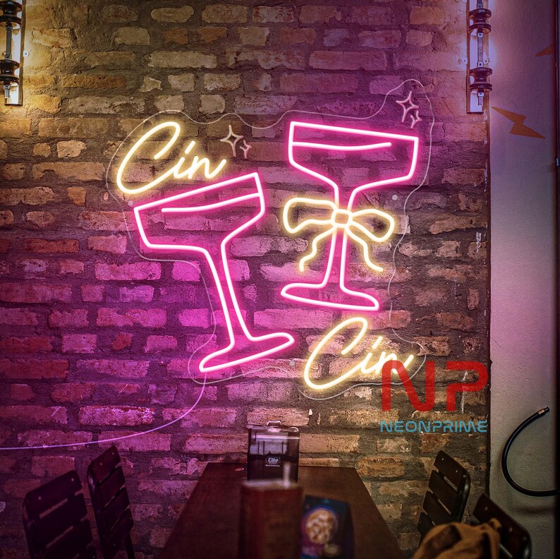 Pu&ograve; includere: Un'insegna al neon con le parole "Cin Cin" e due calici da cocktail. I calici sono delineati in rosa, con il testo in giallo caldo. L'insegna &egrave; montata su un muro di mattoni.
