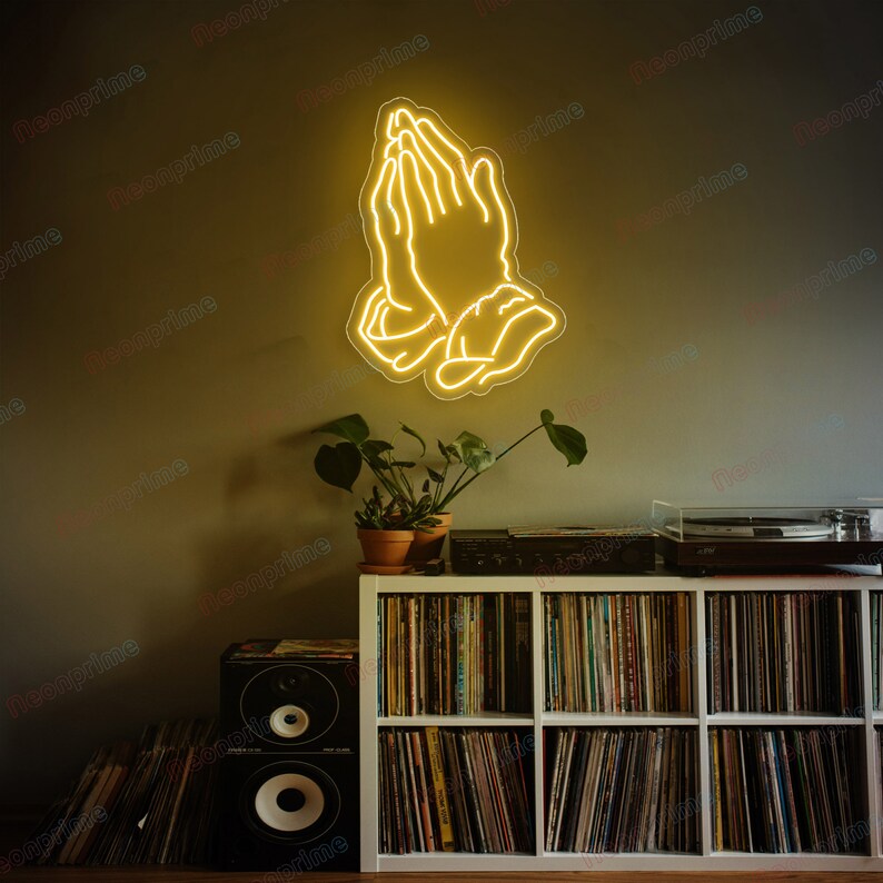 May include: A yellow neon sign depicting two hands clasped in prayer. The sign is mounted on a wall above a white bookshelf filled with vinyl records. A small potted plant sits on the shelf below the sign.