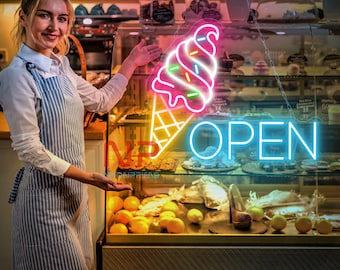May include: A woman in a striped apron gestures towards a neon sign above a display case. The sign shows a colourful ice cream cone and the word "OPEN" in blue. The display case is filled with pastries and fruit, suggesting a bakery or cafe.