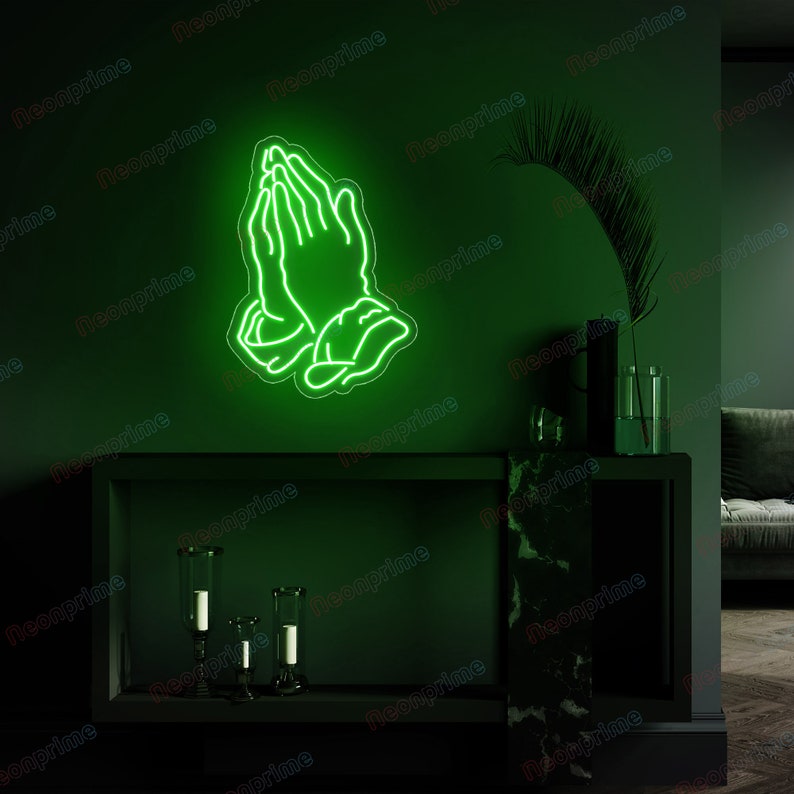 May include: Green neon sign depicting two hands clasped in prayer. The sign is mounted on a dark green wall above a black shelf with decorative candles.