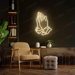 May include: A white neon sign depicting a pair of hands in a praying position. The sign is mounted on a dark wall above a brown armchair and a square ottoman.