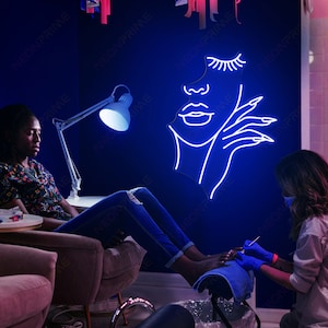 May include: A woman is having a pedicure in a salon. The salon has a blue wall with a neon sign of a woman's face with eyelashes and a hand with long nails.