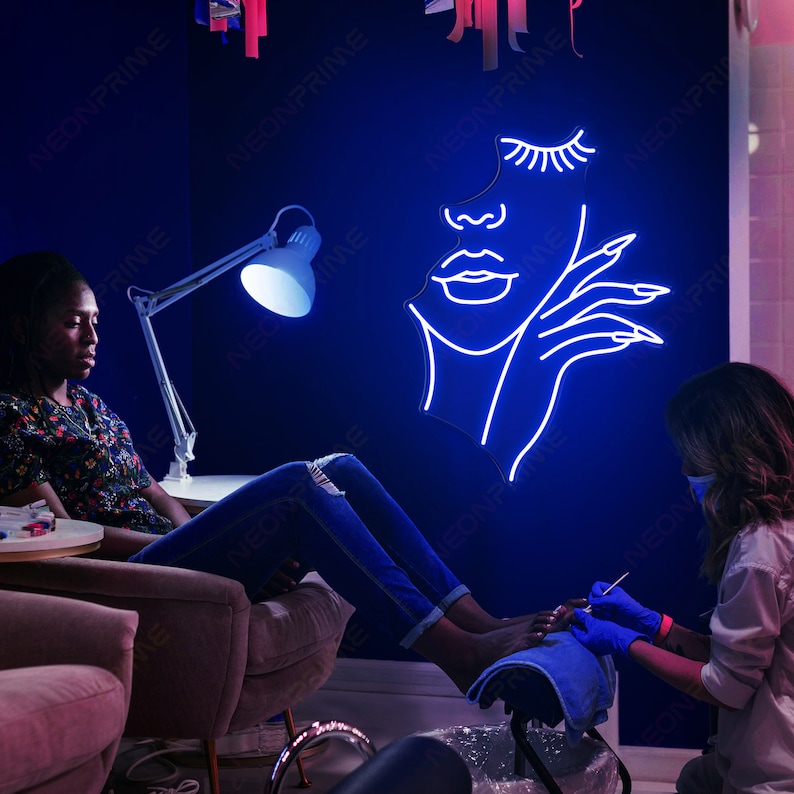 May include: A woman is having a pedicure in a salon. The salon has a blue wall with a neon sign of a woman's face with eyelashes and a hand with long nails.