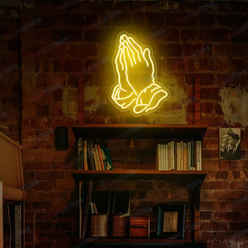 May include: A yellow neon sign depicting two hands clasped in prayer. The sign is mounted on a brick wall above a bookshelf.