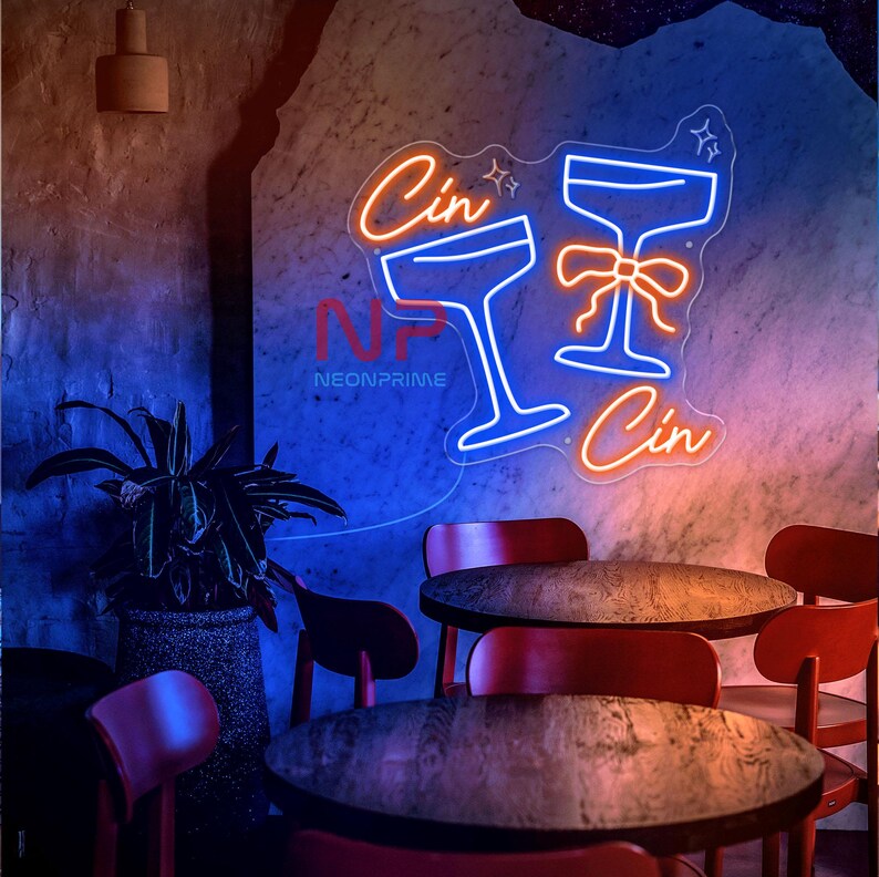 Pu&ograve; includere: Un'insegna al neon con due calici da cocktail e la parola "Cin" in arancione e blu. L'insegna &egrave; montata su una parete strutturata sopra sedie e tavoli rossi. &Egrave; visibile anche il logo "Neonprime".