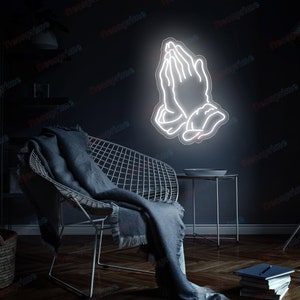 May include: White neon sign of two hands in a praying position against a dark wall. The sign is hanging above a chair with a blanket on it.