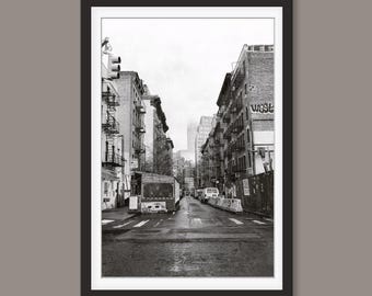 May include: Black and white framed print of a New York City street scene. The image shows a narrow street lined with brick buildings, fire escapes, and a bus. The sky is overcast, and the street is wet.