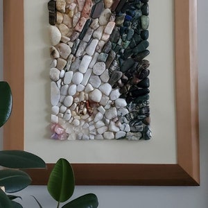 以下が含まれることがあります： カラフルな石と白い石のモザイクでできた額装されたアート作品。石は、風景や抽象的なデザインを思わせるパターンで配置されています。