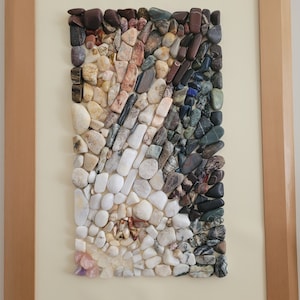 以下が含まれることがあります： 小さなカラフルな石のモザイクでできた額装されたアート作品。石は幾何学的なパターンで配置されており、白、グレー、茶色、黒の石が混ざり合っています。アート作品は白い背景に置かれ、薄い茶色の木製の額縁で囲まれています。