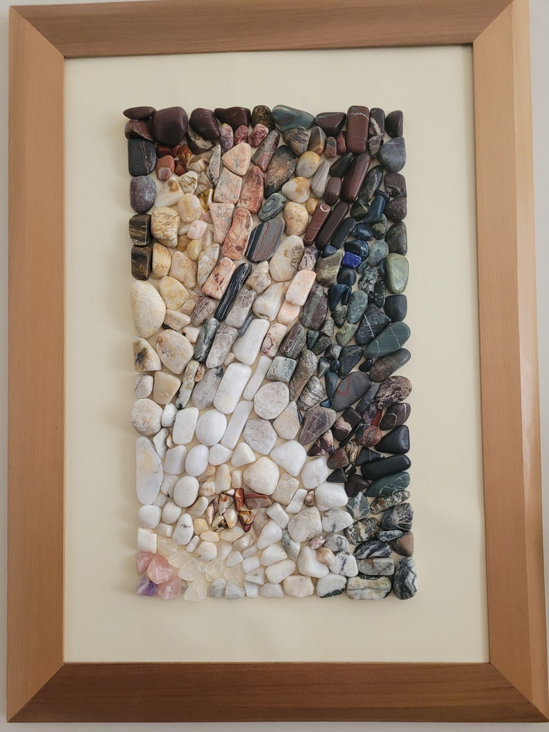 以下が含まれることがあります： 小さなカラフルな石のモザイクでできた額装されたアート作品。石は幾何学的なパターンで配置されており、白、グレー、茶色、黒の石が混ざり合っています。アート作品は白い背景に置かれ、薄い茶色の木製の額縁で囲まれています。