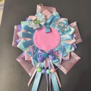 May include: A handmade rosette badge featuring a pink felt circle at the center, surrounded by layers of pastel ribbons in pink, blue, and lavender. Embellishments include bows, beads, and a heart-shaped charm. The badge has a metal loop for hanging.