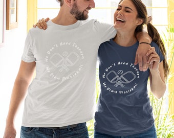 Op de afbeelding: Een stel draagt ​​bijpassende T-shirts met de tekst "We Don't Need Therapy We Play Pickleball" en een afbeelding van twee pickleball-rackets. De man draagt ​​een wit T-shirt en de vrouw draagt ​​een donkerblauw T-shirt.