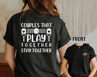 Op de afbeelding: Zwart T-shirt met witte tekst die "Couples that play together stay together" luidt. De tekst wordt vergezeld door een afbeelding van twee pickleball-rackets en een pickleball.