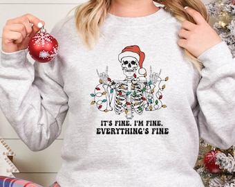 May include: A gray sweatshirt with a skeleton wearing a Santa hat and tangled in Christmas lights. The text "It's fine. I'm fine. Everything's fine" is printed below the skeleton.